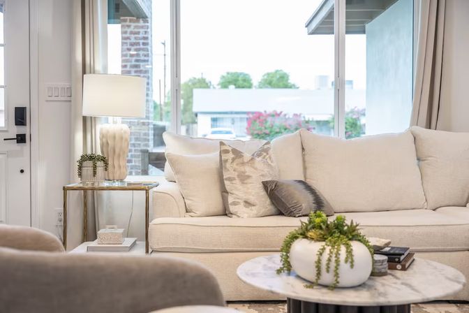 Sunlit modern living room with cream sofa and patterned throw pillows, marble coffee table with a succulent planter, gold side table and lamp, and a large window showing suburban greenery.