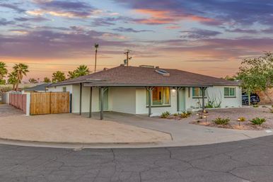 Single-story midcentury ranch home with carport, white brick exterior and xeriscape front yard with palm trees, set against a colorful Southwestern sunset sky.