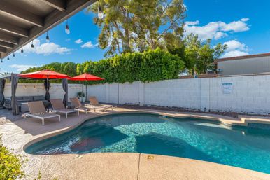 Sunny residential backyard swimming pool with clear blue water, curved concrete deck, four beige lounge chairs under bright red umbrellas, gray cabana with curtains, string lights hanging from a covered patio, and a white block wall with green privacy hedge.