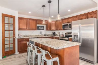 Bright modern home kitchen with a large speckled granite island breakfast bar and three metal stools, warm wood cabinets, stainless steel refrigerator and stove, industrial pendant lights, and a glass-paneled pantry door.