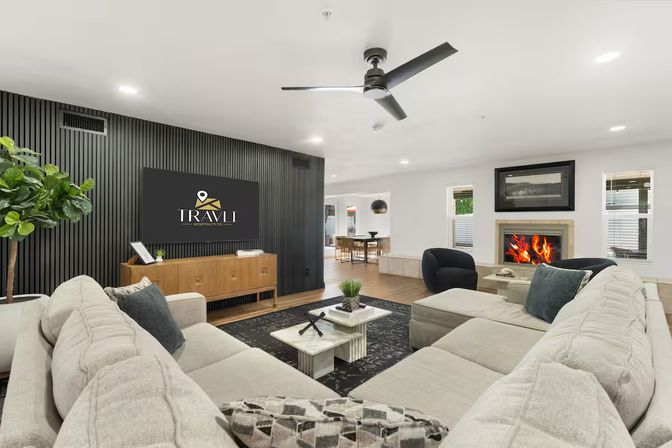 Open-concept modern living room with large beige sectional, black slatted accent wall and TV, wooden media console, ceiling fan, marble coffee table, indoor plant and roaring fireplace