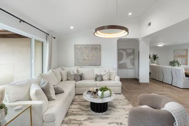 Airy modern open-concept living room with a white L-shaped sectional, marble round coffee table with greenery, circular pendant light, sliding glass doors, neutral rug and herringbone wood floors leading to a dining area.
