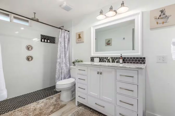 Bright modern white bathroom with marble-top vanity and large mirror, black hexagon tile accents, walk-in shower with patterned curtain and plush rugs.