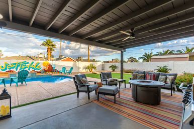 Covered Scottsdale backyard patio with cushioned seating around a round fire pit on a striped rug, ceiling fan, swimming pool with turquoise Adirondack chairs, colorful desert mural on the wall, palm trees and manicured lawn.