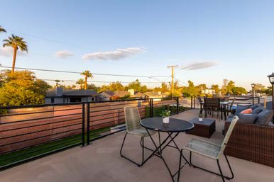Sunlit rooftop patio at golden hour overlooking a palm‑lined suburban neighborhood, featuring a bistro table with potted plant, wicker sofa with cushions, outdoor dining set and metal railing.