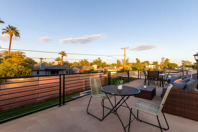 Sunlit rooftop patio at golden hour overlooking a palm‑lined suburban neighborhood, featuring a bistro table with potted plant, wicker sofa with cushions, outdoor dining set and metal railing.