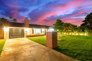 Well-lit modern ranch-style suburban home with striped garage door, brick pillar, lush front lawn and vibrant pink-purple sunset sky