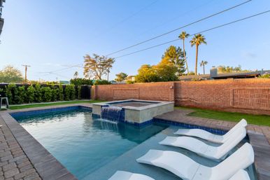 Suburban backyard pool with raised spa waterfall, three white loungers on a tanning ledge, brick privacy wall and palm trees under a clear evening sky.