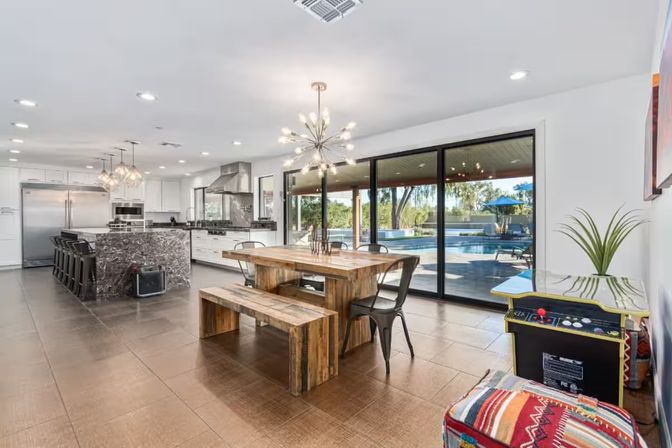 Sunlit modern open-plan kitchen and dining area with marble island and bar stools, rustic wooden table and bench, metal chairs, sputnik chandelier and large sliding glass doors opening to a backyard pool and patio.