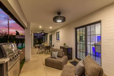 Cozy covered backyard patio at sunset with stainless-steel grill, outdoor dining set, cushioned lounge seating, string lights, and sliding glass doors to the kitchen.