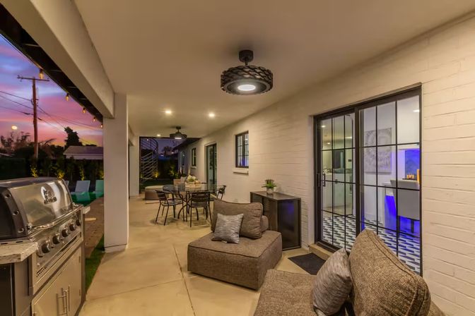 Cozy covered backyard patio at sunset with stainless-steel grill, outdoor dining set, cushioned lounge seating, string lights, and sliding glass doors to the kitchen.