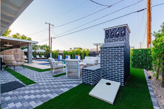 Modern backyard patio with rectangular pool, wicker lounge chairs and umbrellas, black-brick outdoor fireplace, cornhole board on artificial turf, paver walkway and hedged privacy fence.