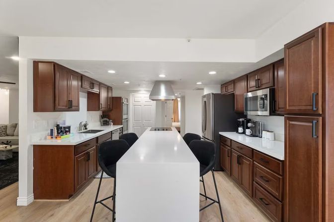 Bright modern open-plan kitchen with a long white island and four black bar stools, stainless steel range hood and appliances, dark wood cabinets, white countertops and light hardwood floors.