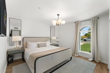 Sunlit modern bedroom with a king bed in neutral linens, matching nightstands and glass lamps, contemporary chandelier, and an arched window with curtains overlooking a green lawn and quiet suburban street.