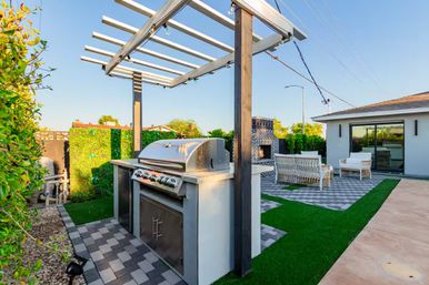 Modern backyard patio with built-in stainless steel grill under a white pergola with string lights, paver flooring, brick outdoor fireplace and white lounge seating on artificial turf.