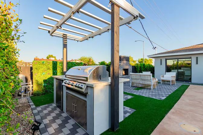 Modern backyard patio with built-in stainless steel grill under a white pergola with string lights, paver flooring, brick outdoor fireplace and white lounge seating on artificial turf.