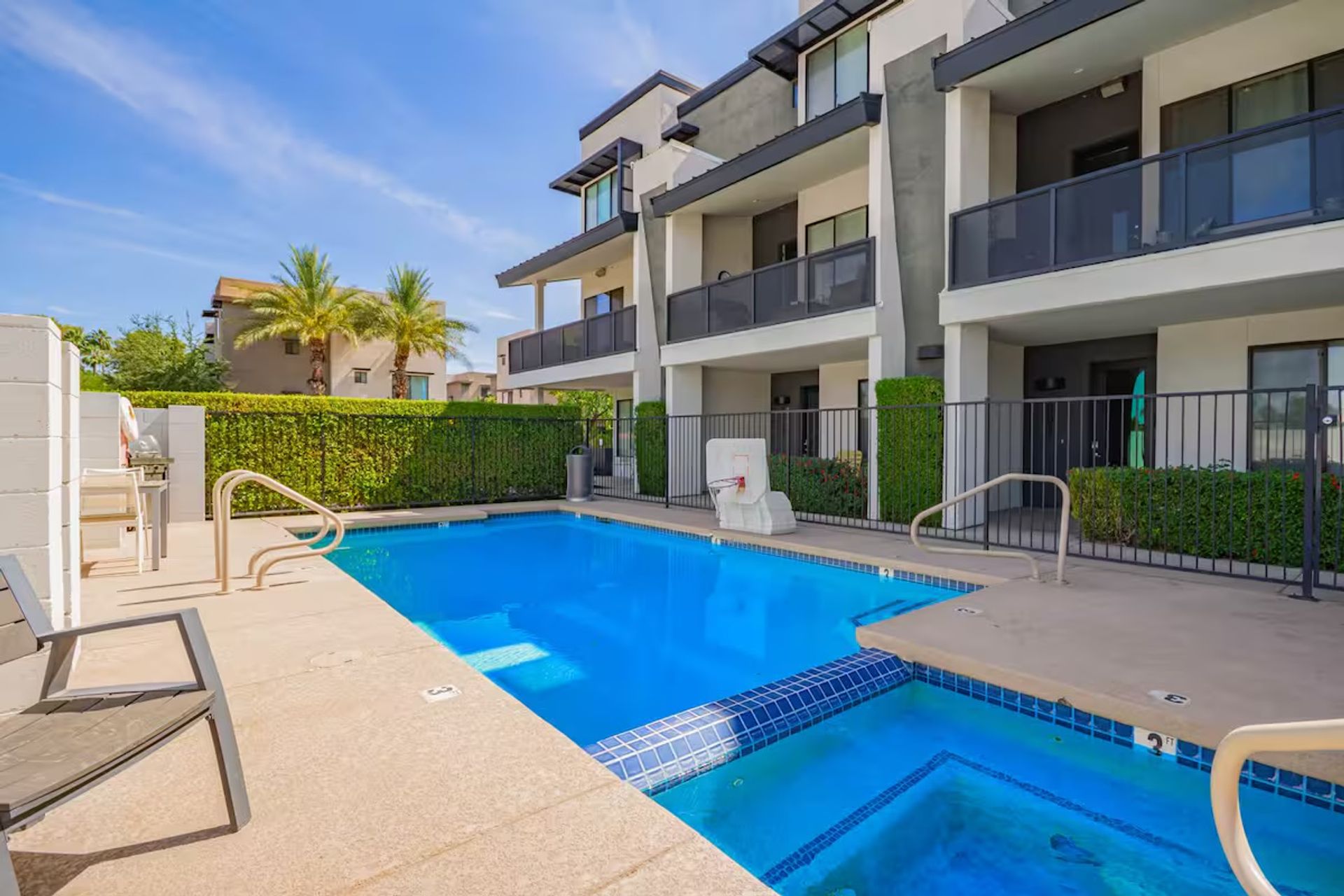 Sparkling blue pool and attached hot tub at a sunny modern apartment complex with balcony units, palm trees, lounge chairs and a gated patio