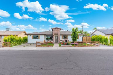 Single-story ranch-style suburban home with brick entry tower, white stucco exterior, wooden cross-fencing, manicured lawn, colorful flowerbeds and paved driveway under a bright blue sky