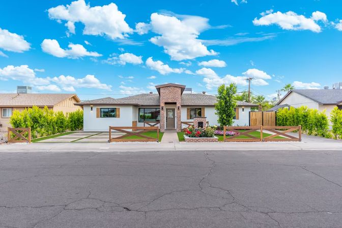 Single-story ranch-style suburban home with brick entry tower, white stucco exterior, wooden cross-fencing, manicured lawn, colorful flowerbeds and paved driveway under a bright blue sky