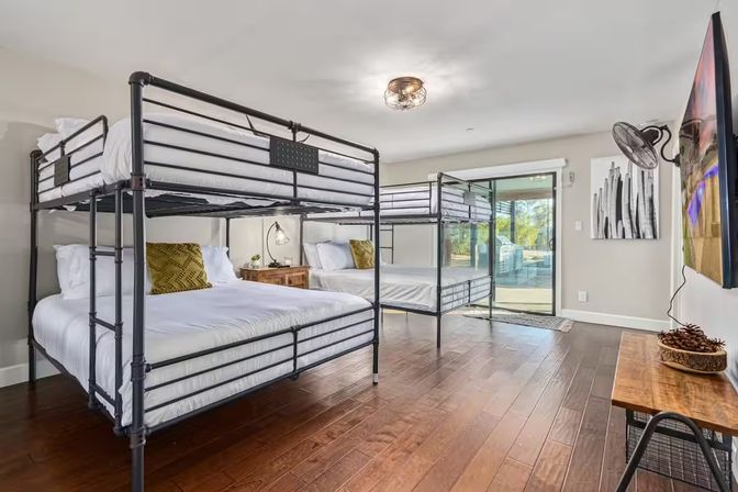 Bright modern vacation-rental bedroom with metal double-over-double bunk beds, hardwood floors, nightstand and sliding glass door to a patio.