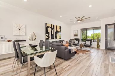 Bright modern open-concept living and dining room with gray sectional, glass dining table, wood-look tile floors, ceiling fan, and large window overlooking greenery.