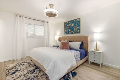 Bright modern bedroom with a tufted beige headboard, navy-and-white bedding and a blush accent pillow, floral wall art, matching nightstands with lamps, white curtains, patterned rug and light wood floors.