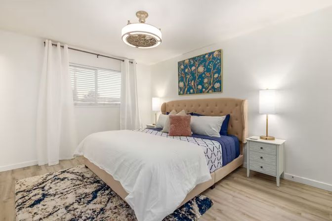 Bright modern bedroom with a tufted beige headboard, navy-and-white bedding and a blush accent pillow, floral wall art, matching nightstands with lamps, white curtains, patterned rug and light wood floors.