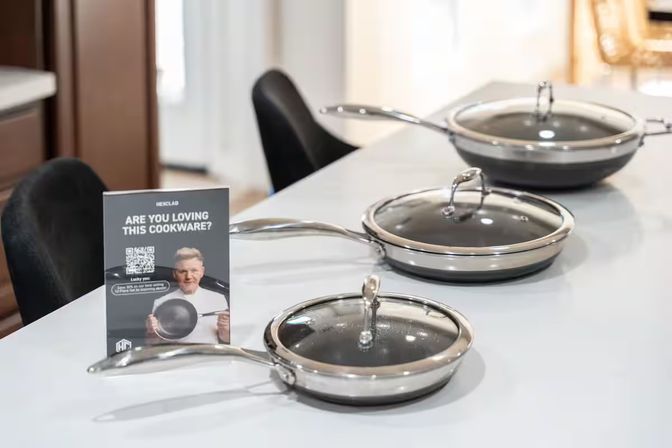 Three gleaming stainless-steel nonstick frying pans with glass lids displayed on a white kitchen island beside a promotional sign.