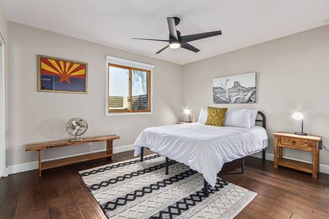 Modern bright bedroom with white linens on a metal bed frame, black-and-white patterned rug on hardwood floors, ceiling fan, wooden bench and nightstands, and Arizona-inspired wall art