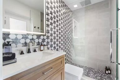 Modern home bathroom with wood vanity and white marble countertop, black matte faucets, illuminated mirror, geometric black-and-white hexagon tile accent wall and mosaic shower floor, glass walk-in shower