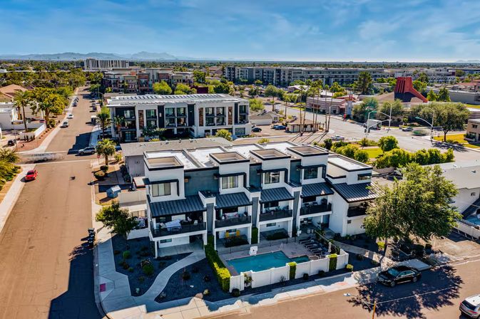 Aerial view of a modern white townhome complex with rooftop terraces and a central turquoise pool in a sunny suburban neighborhood, tree-lined streets, low-rise apartments and distant mountains under a blue sky.