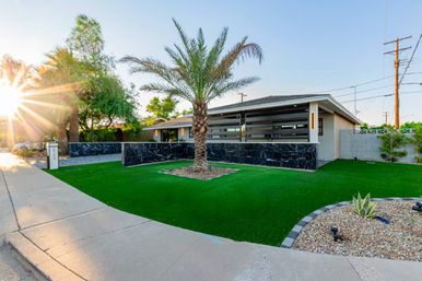 Mid-century modern suburban home at sunset with a central palm tree, black marble low wall, bright green artificial turf lawn, gravel rock garden with agave, sidewalk and sunburst glow