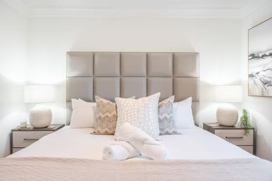Contemporary master bedroom with tufted gray headboard, crisp white bedding, patterned neutral throw pillows, rolled towels on the bed, and matching nightstands with woven lamps and soft ambient lighting.