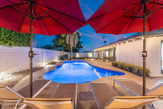 Inviting twilight backyard with an illuminated kidney-shaped swimming pool, red patio umbrellas, lounge chairs, string lights along a single-story home and a palm tree.