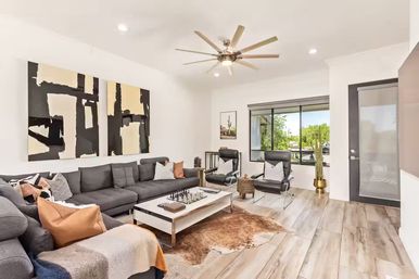Sunlit modern living room with a long gray sectional, abstract black-and-beige wall art, white coffee table with a chess set, two black leather lounge chairs, a large ceiling fan, wood-look tile floors and a big window showing green yard views.