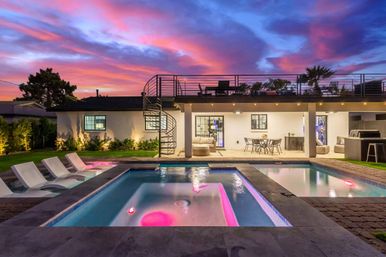 Luxury modern backyard at sunset with vivid pink and purple sky, illuminated rectangular pool and spa with pink LED lights, built-in loungers, outdoor dining and lounge beneath a rooftop deck accessed by a spiral staircase.