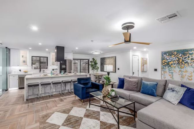 Modern open-concept living room and kitchen with gray sectional, blue accent chair, glass coffee table, long island with bar stools, wood-look herringbone tile floor, ceiling fan and abstract wall art.