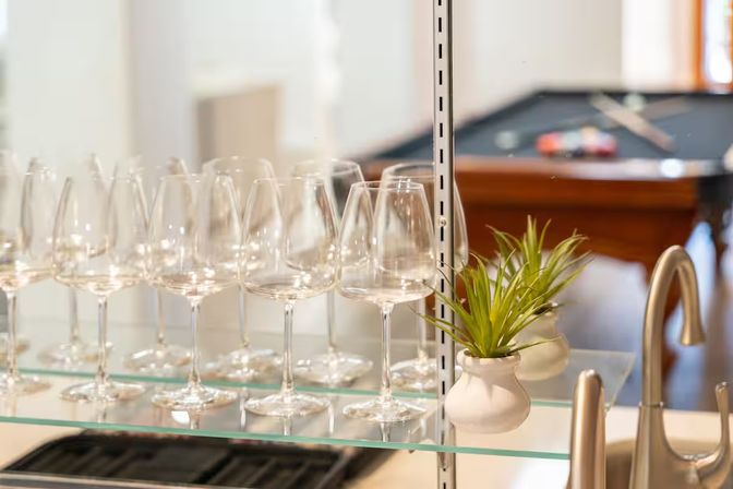 Row of sparkling wine glasses on a glass kitchen bar shelf with a small potted succulent and a blurred pool table in the background.