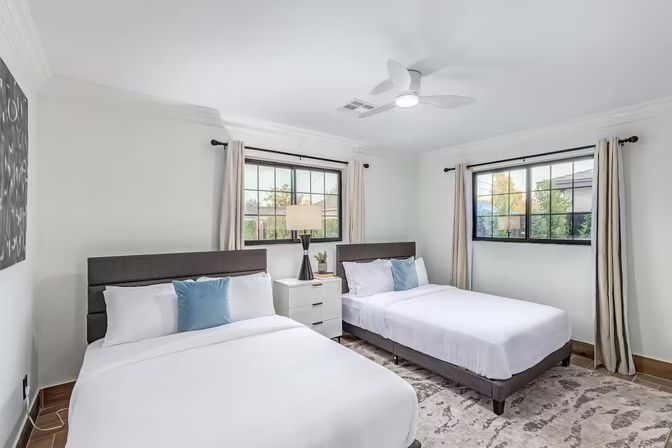 Bright contemporary guest bedroom with two queen beds, crisp white linens and blue accent pillows, gray headboards, shared white nightstand with lamp, black-framed windows with beige curtains, ceiling fan and patterned area rug