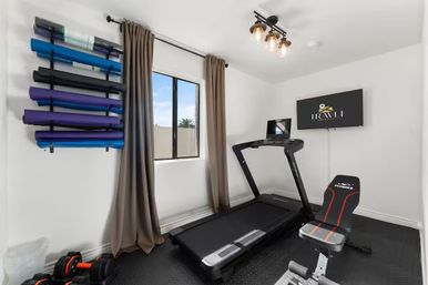 Compact bright home workout room with treadmill, adjustable bench, dumbbells, wall rack of blue and purple yoga mats, wall-mounted TV, window with curtains and overhead lighting.