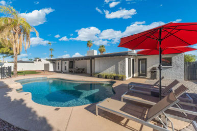Sunny backyard with curved blue swimming pool, loungers under a red umbrella, covered patio dining area, palm trees and single-story stucco home