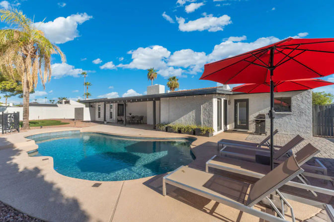 Sunny backyard with curved blue swimming pool, loungers under a red umbrella, covered patio dining area, palm trees and single-story stucco home
