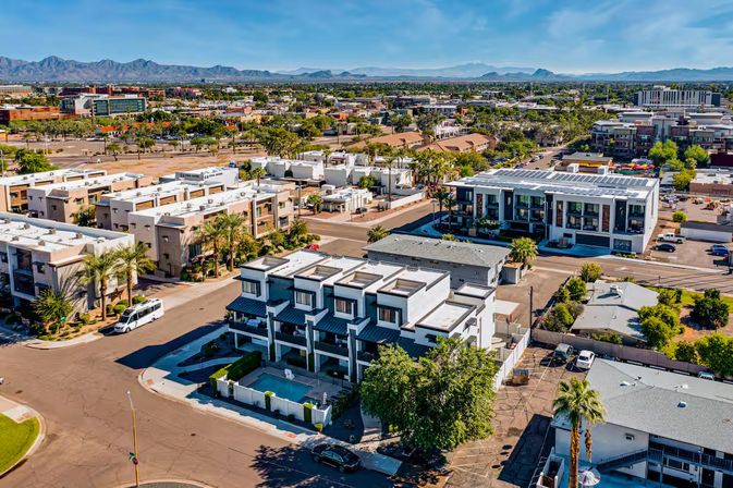 Aerial view of a sun-soaked Southwestern urban neighborhood with modern white townhouses, palm trees, rooftop solar panels, parking areas and a distant mountain range under a blue sky.