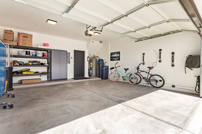 Bright two-car residential garage with open door, concrete floor, organized shelving and cabinets, wall-mounted bike racks holding two bicycles, and utility equipment.