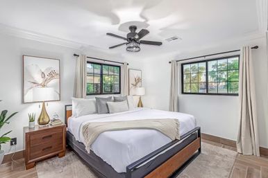 Bright, modern bedroom interior with a king-size bed in white linens, gray pillows and beige throw, black metal ceiling fan, two black-framed windows with leafy views, wooden nightstands with gold lamps and botanical artwork, neutral curtains and hardwood floors.