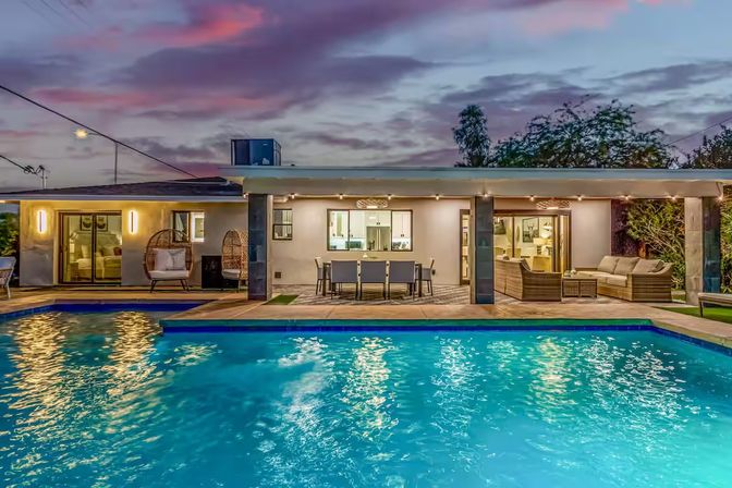 Twilight backyard of a modern single-story home featuring a glowing blue swimming pool, covered patio with outdoor dining table, wicker lounge seating and hanging egg chairs, and warm interior lights visible through sliding glass doors.