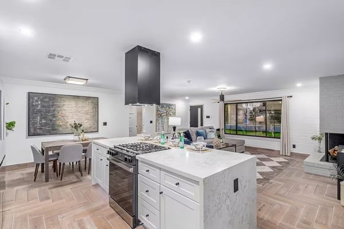 Bright modern open-concept kitchen and living area with marble-topped island and stainless gas range, white cabinetry, dining table, cozy seating, large picture window, fireplace, and wood-look tile floors.