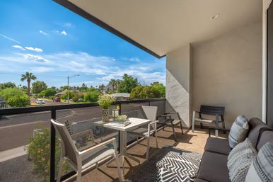 Sunny modern balcony with small dining table, lounge sofa and chairs, potted flowers, overlooking a palm-lined suburban street under a bright blue sky.