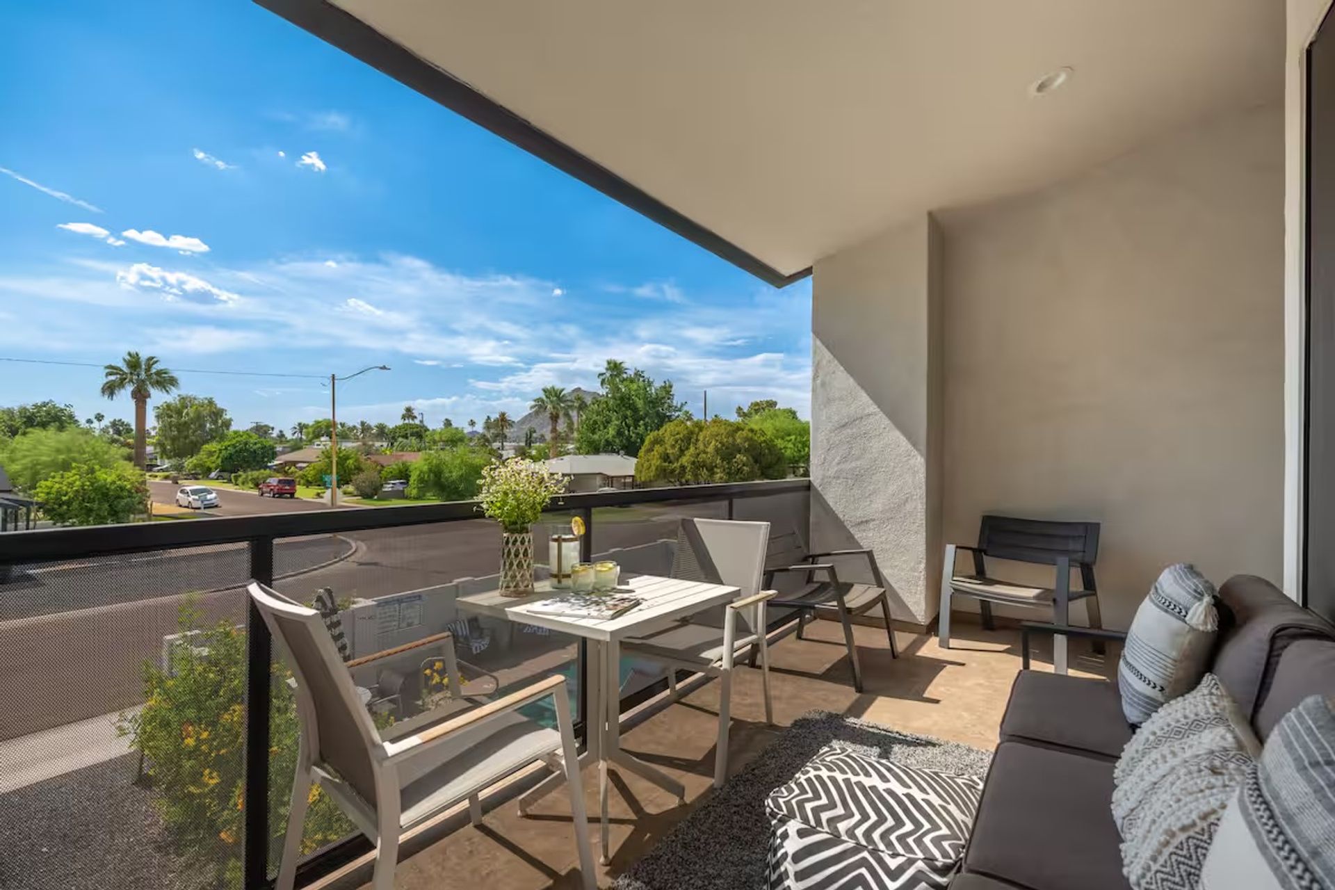 Sunny modern balcony with small dining table, lounge sofa and chairs, potted flowers, overlooking a palm-lined suburban street under a bright blue sky.