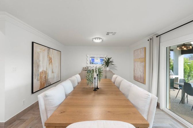 Airy modern dining room with a long oak table and white upholstered chairs, neutral abstract wall art, a greenery centerpiece and potted plant on a sideboard, minimalist ceiling light, and a sliding glass door opening to a patio.